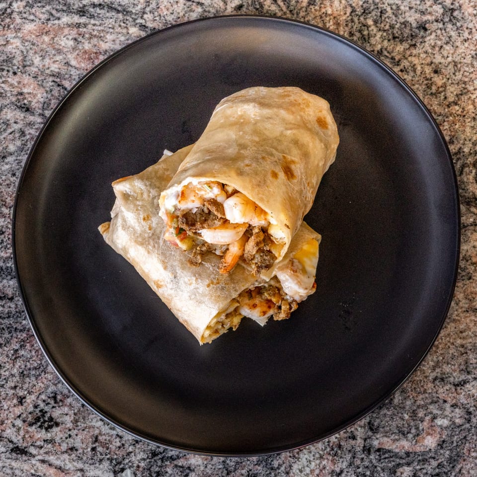 Two large burritos, filled with meat and shrimp, on a black plate atop a granite counter.
