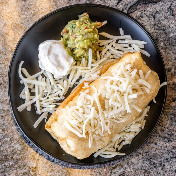 Golden-fried chimichanga topped with shredded cheese, served with guacamole and sour cream on a black plate.