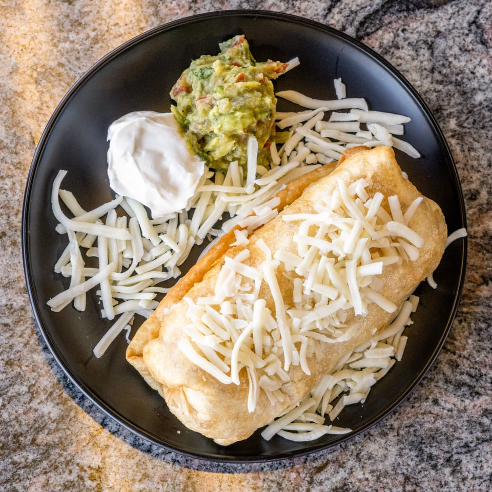 Golden-fried chimichanga topped with shredded cheese, served with guacamole and sour cream on a black plate.