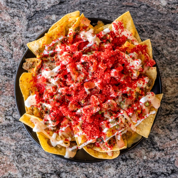 A black plate of loaded nachos with red seasoning, white sauce, and pico de gallo.