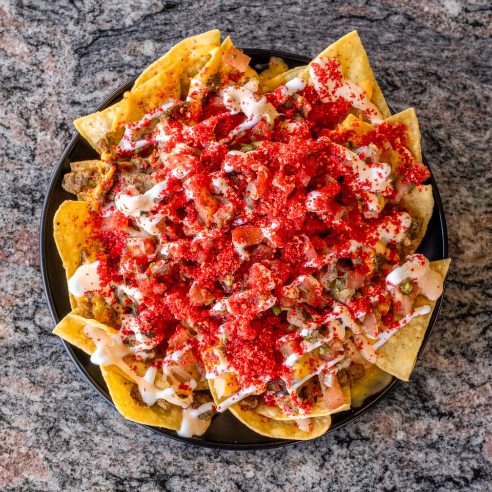 A black plate of loaded nachos with red seasoning, white sauce, and pico de gallo.