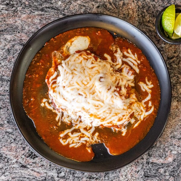 A black plate holds a dish covered in red sauce and melted white cheese, with lime wedges nearby.