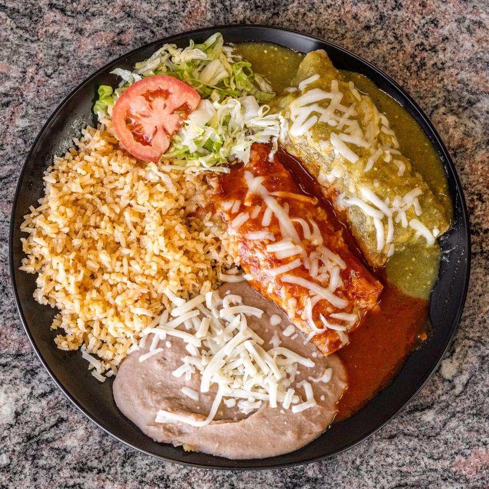 A black plate of Mexican food with rice, beans, two enchiladas, lettuce, and tomato.