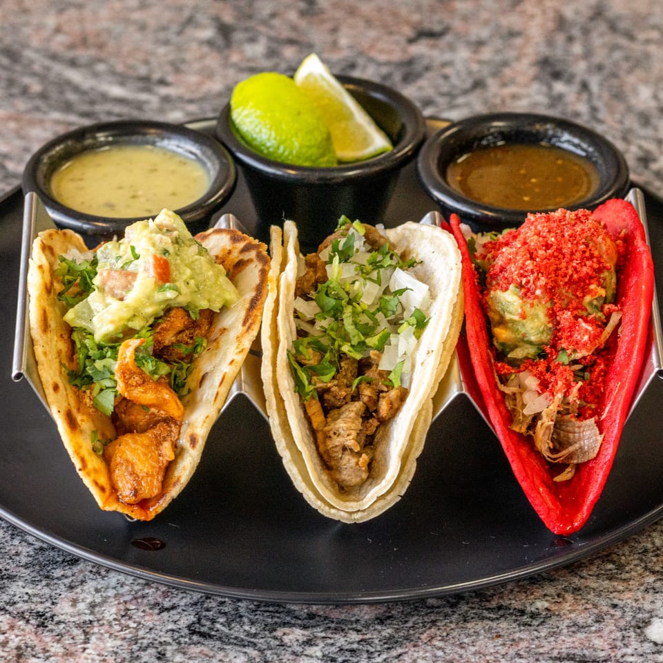 Three vibrant tacos with various fillings, sauces, and lime wedges on a black plate.
