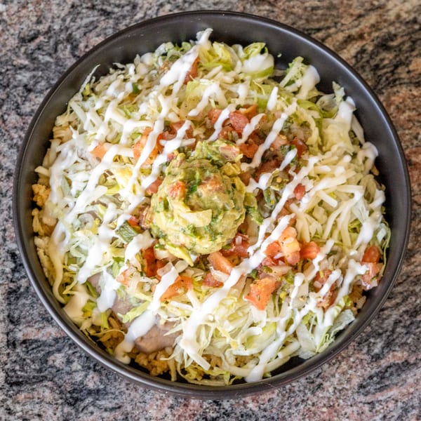 A black bowl filled with a burrito bowl, topped with lettuce, cheese, pico de gallo, guacamole, and sour cream.