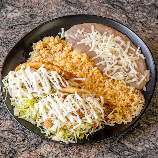 Two crispy tacos with lettuce, sour cream, rice, and refried beans with cheese on a black plate.